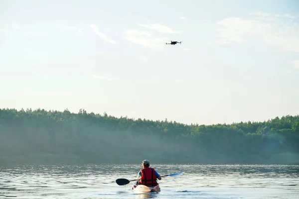 男は湖のカヤックに乗り無人機が彼の上を飛んでいます — ストック写真
