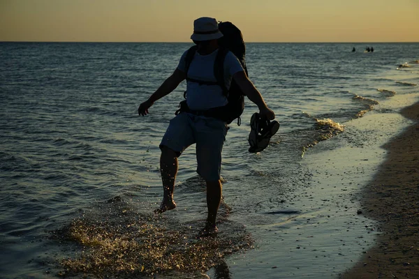 Şort, Panama tişörtü ve gözlüklü, büyük sırt çantalı bir turist gün batımında deniz kıyısında ıslak kum boyunca yürür..