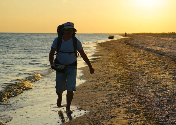 Şort, Panama tişörtü ve gözlüklü, büyük sırt çantalı bir turist gün batımında deniz kıyısında ıslak kum boyunca yürür..