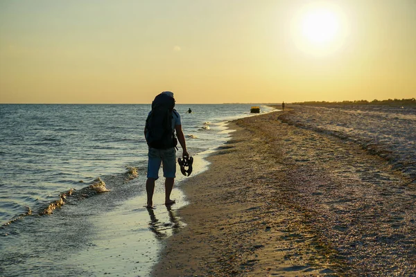 Sırt çantalı bir adam kumsalda yalınayak yürüyor, elinde sandaletleri var.