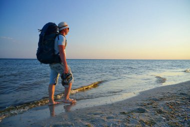 Sırt çantalı bir adam kumsalda yalınayak yürüyor, elinde sandaletleri var.