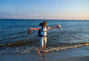 Sırt çantalı bir adam deniz kıyısında yalınayak durur ve bir elinde Panama, diğer elinde sandalet tutar.