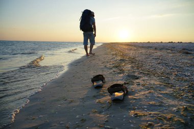 Deniz kenarında ıslak kumda sandaletler. Adam sahil boyunca sırt çantasıyla yürüyor. Akşam güneşinin ışınları ıslak kuma yansıyor.