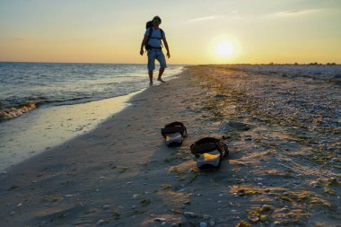 Deniz kenarında ıslak kumda sandaletler. Adam sahil boyunca sırt çantasıyla yürüyor. Akşam güneşinin ışınları ıslak kuma yansıyor.