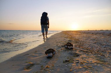 Deniz kenarında ıslak kumda sandaletler. Adam sahil boyunca sırt çantasıyla yürüyor. Akşam güneşinin ışınları ıslak kuma yansıyor.
