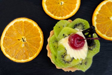 Yanında kivili ve kremalı pasta, portakal dilimleri var. Siyah arkaplanda