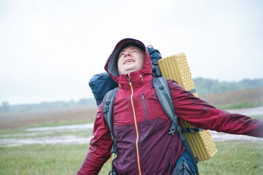 Kırmızı ceketli, sırt çantalı ve seyahat minderli bir turist yağmurda duruyor.