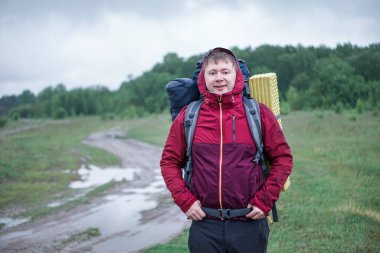 Turist yürüyüşçüsü yağmurda kırmızı ceketli bir sırt çantasıyla yolda yürüyor.