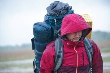 Kırmızı ceketli, sırt çantalı ve seyahat minderli bir turist yağmurda duruyor.