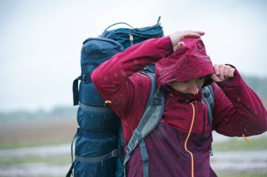 Kırmızı ceketli, sırt çantalı ve seyahat minderli bir turist yağmurda duruyor.