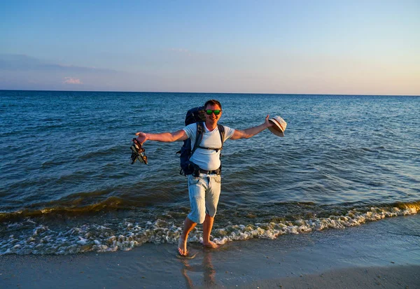 Sırt çantalı bir adam deniz kıyısında yalınayak durur ve bir elinde Panama, diğer elinde sandalet tutar.