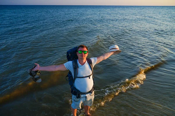 Sırt çantalı bir adam deniz kıyısında yalınayak durur ve bir elinde Panama, diğer elinde sandalet tutar.