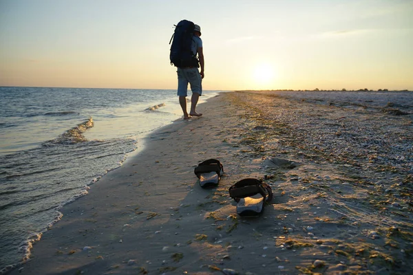 Deniz kenarında ıslak kumda sandaletler. Adam sahil boyunca sırt çantasıyla yürüyor. Akşam güneşinin ışınları ıslak kuma yansıyor.