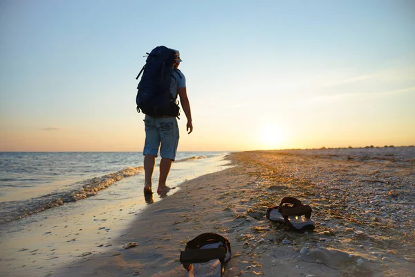 Deniz kenarında ıslak kumda sandaletler. Adam sahil boyunca sırt çantasıyla yürüyor. Akşam güneşinin ışınları ıslak kuma yansıyor.