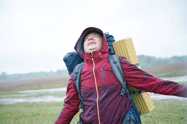 Kırmızı ceketli, sırt çantalı ve seyahat minderli bir turist yağmurda duruyor.