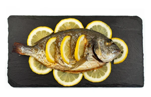 Fırında pişirilmiş Dorado balığı ve limon siyah bir taş tahtada yatıyor.