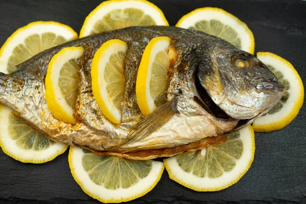 Fırında pişirilmiş Dorado balığı ve limon siyah bir taş tahtada yatıyor.