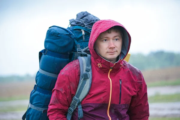 Kırmızı ceketli, sırt çantalı ve seyahat minderli bir turist yağmurda duruyor.