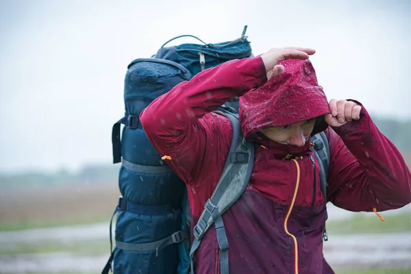 Kırmızı ceketli, sırt çantalı ve seyahat minderli bir turist yağmurda duruyor.