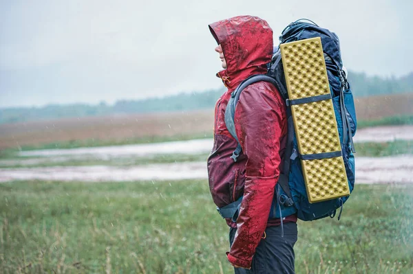 Kırmızı ceketli, sırt çantalı ve seyahat minderli bir turist yağmurda duruyor.