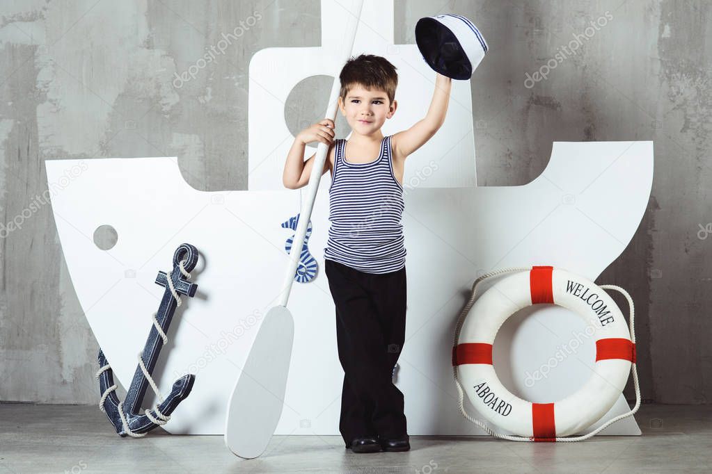 Cabin boy in striped t-shirt waving sailor cap in front of stylized ship, studio shot