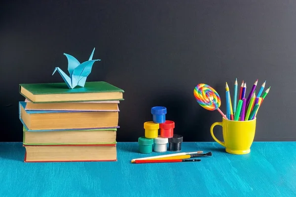 Pupils workplace. Books stationery paint gouache and origami crane on blue table on background black chalkboard Concept Education and Back to school