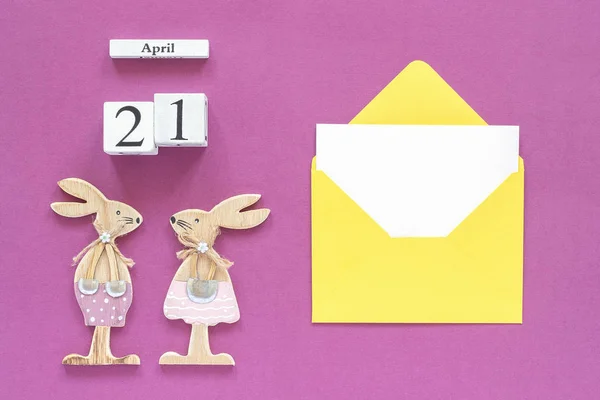 Kalender 21. April, Paar hölzerne Osterhasen, gelber Umschlag mit leerer Karte auf lila Papier Hintergrundkonzept katholische Osterattrappe für Schriftzug, Text oder Ihre Designvorlage von oben — Stockfoto