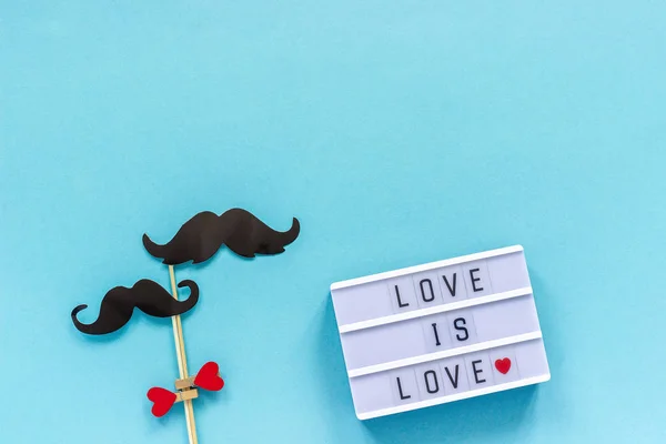 Paar Papier Schnurrbart Requisiten auf Stock und Leuchtkasten mit Text Liebe ist Liebe auf blauem Hintergrund. Begriff Homosexualität Homosexuelle Liebe. Internationaler Schwulentag oder Valentinstag Top-Ansicht Grußkarten-Vorlage — Stockfoto