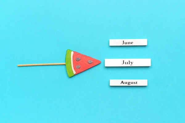Calendario in legno mesi estivi luglio, giugno, agosto e anguria lecca-lecca su bastone su sfondo blu. Concetto vacanza, vacanze o Ciao luglio. Modello creativo vista dall'alto — Foto Stock