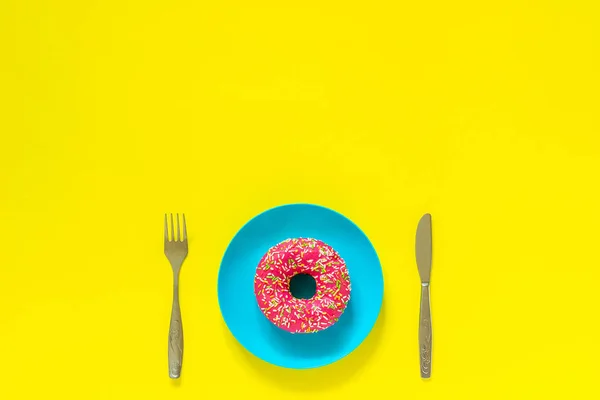 Donut rosa sobre plato azul y tenedor de cuchillo de cubiertos sobre fondo amarillo . — Foto de Stock