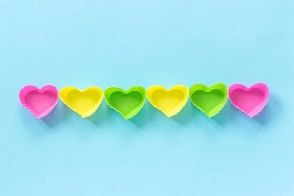 Placa de moldes em forma de coração de silicone colorido para assar cupcakes alinhados em linha no fundo de papel azul. Modelo para lettering, texto ou seu design Vista superior, espaço de cópia — Fotografia de Stock