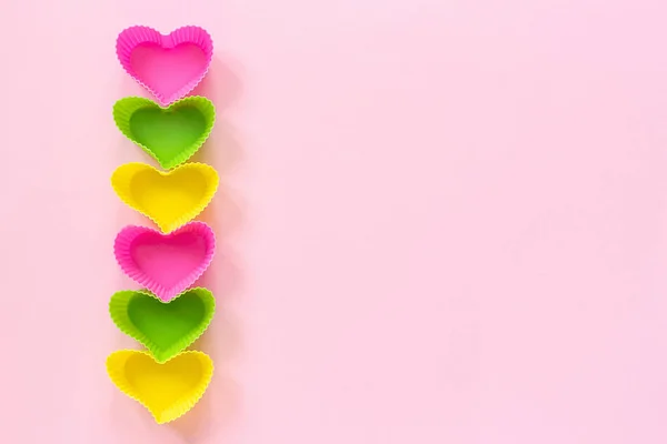 Plato de moldes en forma de corazón de silicona de color para hornear magdalenas forradas en el borde izquierdo de la fila sobre fondo de papel rosa. Plantilla para letras, texto o tu diseño Vista superior, espacio para copiar — Foto de Stock