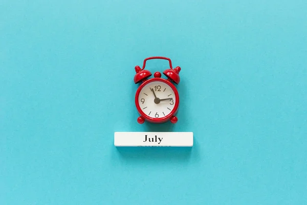 Wooden calendar summer month July and red alarm clock on blue paper background. Concept Hello July or Good bye July Creative Top view Flat Lay Minimal style — Stock Photo, Image