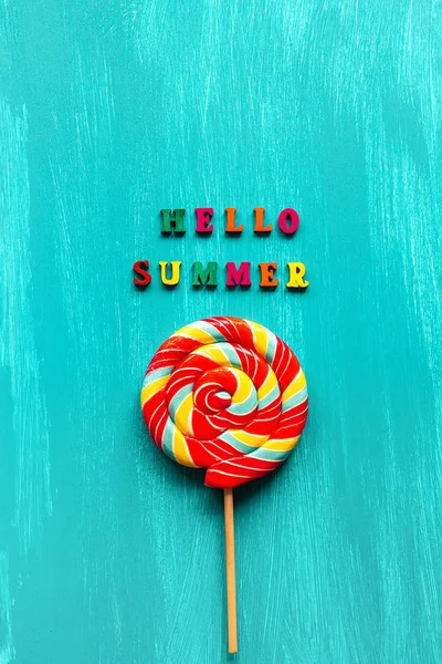 Hello summer text from colorful letters and spiral multi-colored round lollipop on stick on wooden blue background. Concept Creative Top view Template Greeting card — Stock Photo, Image