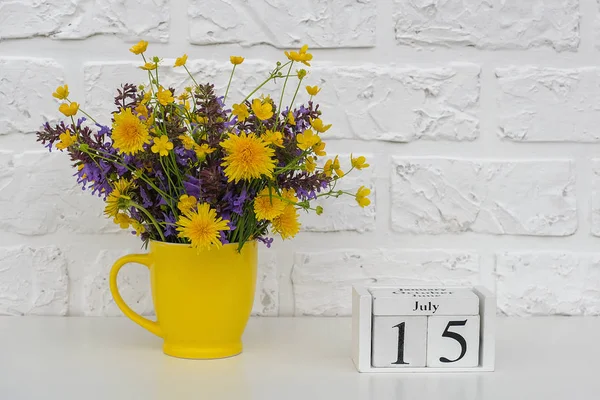 Calendario de cubos de madera 15 de julio y copa amarilla con flores de colores brillantes contra la pared de ladrillo blanco. Plantilla calendario fecha para su diseño Copiar espacio —  Fotos de Stock