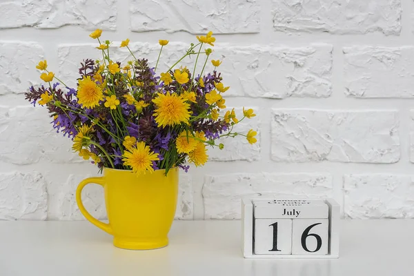 Calendario de cubos de madera 16 de julio y copa amarilla con flores de colores brillantes contra la pared de ladrillo blanco. Plantilla calendario fecha para su diseño Copiar espacio —  Fotos de Stock