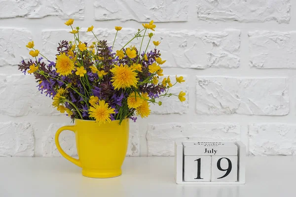 Calendario de cubos de madera 19 de julio y copa amarilla con flores de colores brillantes contra la pared de ladrillo blanco. Plantilla calendario fecha para su diseño Copiar espacio —  Fotos de Stock