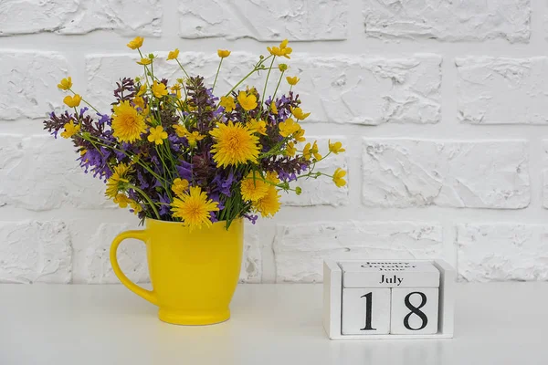 Calendario de cubos de madera 18 de julio y copa amarilla con flores de colores brillantes contra la pared de ladrillo blanco. Plantilla calendario fecha para su diseño Copiar espacio —  Fotos de Stock