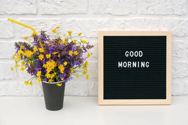 Texto Buenos días en tablero de letras negro y ramo de flores de colores en taza de café de papel negro con paja cóctel en la pared de ladrillo blanco de fondo. Plantilla creativa, Mock up Concept — Foto de Stock