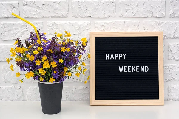 happy weekend words on black letter board and bouquet of yellow dandelions flowers on table against white brick wall. Concept Happy Monday. Template for postcard