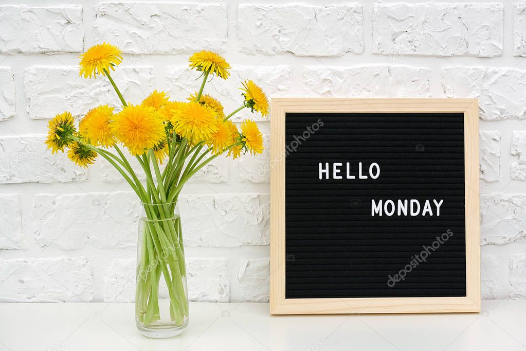 Hello Monday words on black letter board and bouquet of yellow dandelions flowers on table against white brick wall. Concept Happy Monday. Template for postcard