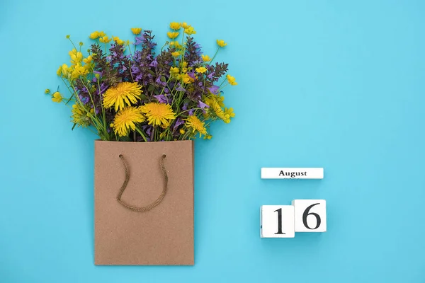 木製の立方体カレンダー8月16日と青い背景にクラフトパッケージでフィールドカラフルな素朴な花。グリーティングカードフラットレイコンセプトこんにちは8月テンプレートテキストとデザインのためのコピースペース付き — ストック写真