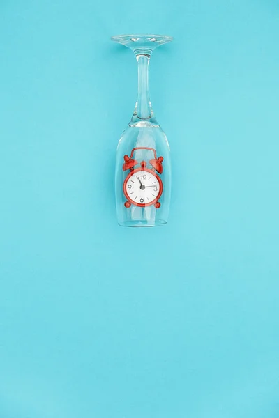 Wine glass with red alarm clock inside on blue background with copy space for text. Creative concept drink time and wine o'clock. Top view Flat lay. Template for invitation, text, design