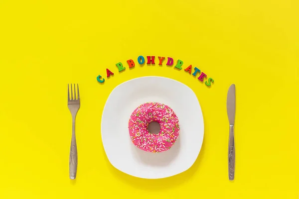 Text carbohydrates, pink donut on plate and cutlery table knife fork . Concept unhealthy diet