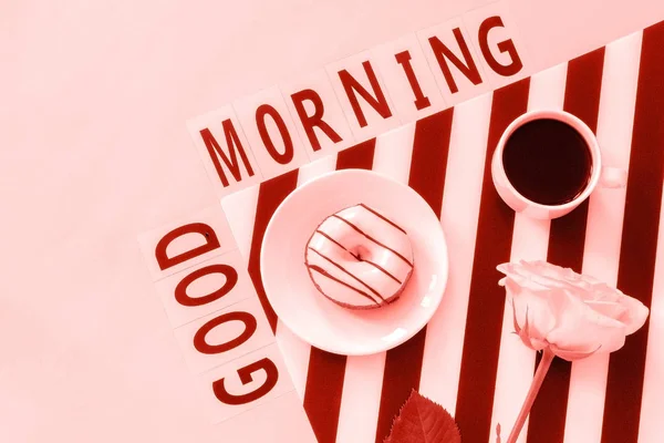 Text Guten Morgen, Kaffee, Donut, Rosenblume auf Serviette und rosa Hintergrund. flache Lay Draufsicht Konzept stilvoller Arbeitsplatz — Stockfoto
