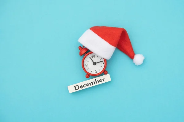 Wooden calendar winter month December, red alarm clock and Santa hat on blue paper background. Concept Hello December, xmas. Creative Top view Flat Lay — Stock Photo, Image