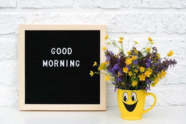 Κείμενο καλό πρωί σε μαύρο γράμμα πλακέτα και μπουκέτο χρωματιστά λουλούδια σε κίτρινο Κύπελλο με ζωγραφισμένο αστείο πρόσωπο στο τραπέζι εναντίον λευκό τοίχο τούβλο. Δημιουργικό πρότυπο, καλλιτεχνικός σχεδιασμός ευτυχισμένο το πρωί — Φωτογραφία Αρχείου