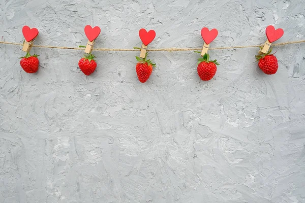 Girlande aus roten reifen Erdbeeren, die an einer hölzernen Wäscheklammer hängen, mit Herzen auf Seilschnur, die sich am oberen Rand auf grauem Hintergrund befinden. Kopierraum kreative Vorlage für Text, Design, Postkarte oder Einladung — Stockfoto