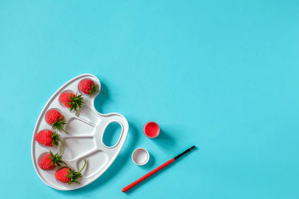 Fresas rojas maduras en una paleta artística, pincel y gouache sobre fondo azul con espacio para copiar. Concepto creativo pintura de colores de verano. Plantilla de laico plano para invitación de postales de diseño — Foto de Stock