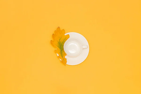 Taza blanca vacía con platillo y hoja de roble de otoño en el centro sobre fondo amarillo. Copiar espacio Flat lay Maquillaje Plantilla para su diseño — Foto de Stock